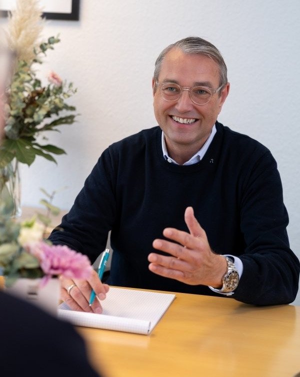 Mann sitz am Tisch, erklärt mit freundlichem Lächeln seiner gegenüber sitzenden Person
